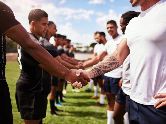 Zwei Gruppen von Rugby-Spielern schütteln sich die Hände auf einem grünen Spielfeld unter blauem Himmel.
