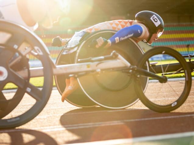 Nahaufnahme zweier Rennrollstühle auf der Laufbahn, während Sportler im Wettkampf um die Wette fahren.