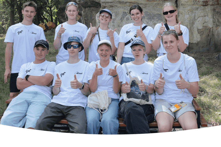 Gruppe von Jugendlichen in weißen T-Shirts zeigt Daumen hoch, sitzend auf einer Bank vor Felsen im Freien.