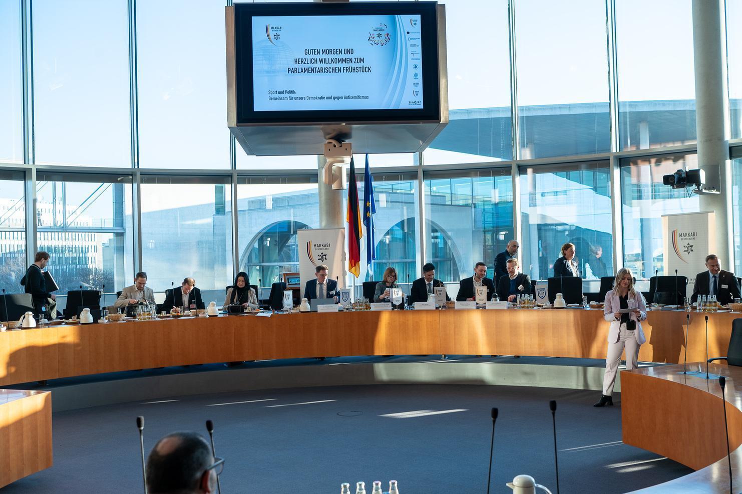 Konferenzraum mit einem langen Tisch, an dem zahlreiche Personen sitzen, und einem Bildschirm im Hintergrund.