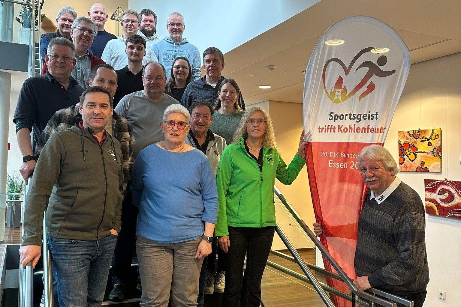 Gruppenfoto von 20 Personen auf einer Treppe, umgeben von einem Banner mit der Aufschrift „Sportgeist trifft Kohlenfeuer“.