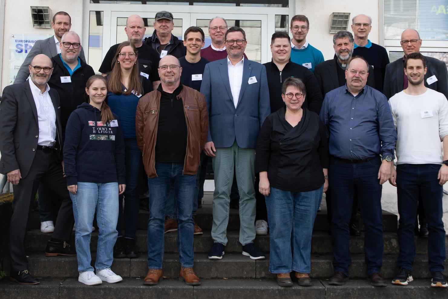 Gruppenfoto von 24 Personen vor einem Gebäude, alle lächeln und tragen Namensschilder.
