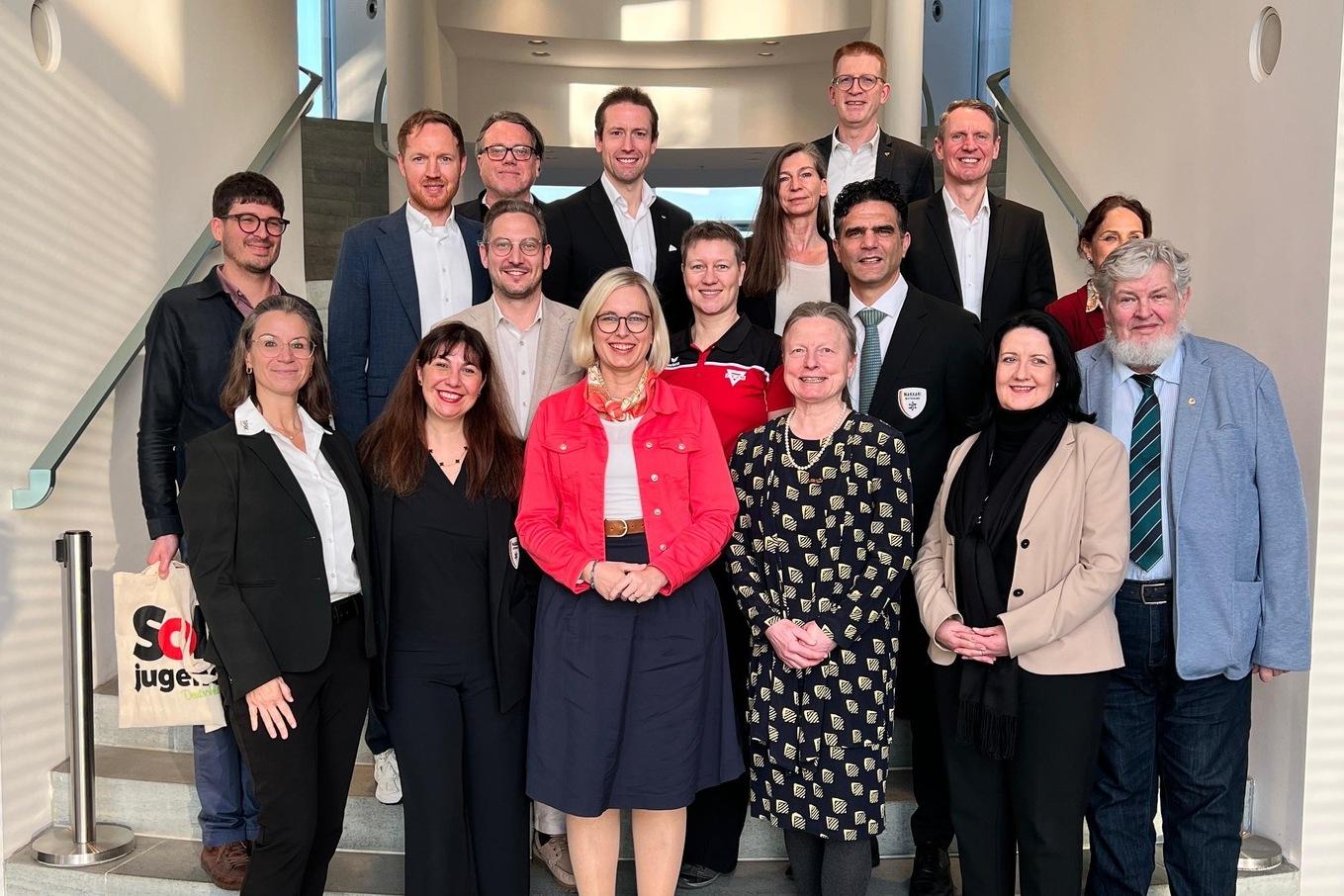 Gruppe von 15 Personen in formeller Kleidung auf einer Treppe, lächelnd und in lockerer Pose.