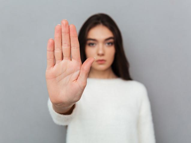 Junge Frau in weißem Pullover hält ihre Hand mit ausgestreckten Fingern in die Kamera. Hintergrund in Grau.