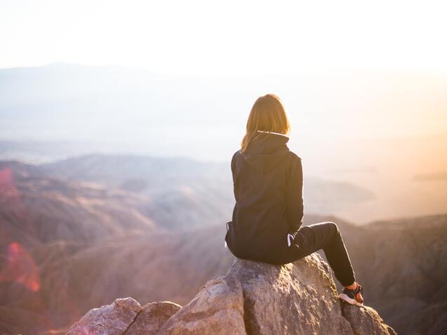 Frau sitzt auf einem Felsen und blickt auf eine weite, bergige Landschaft bei Sonnenaufgang.